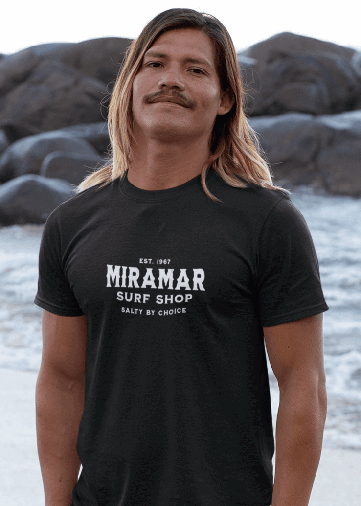 A young man stands on the beach wearing a black Miramar t-shirt. The apparel is a graphic t-shirt designed for men, available in various sizes including large and XXL. This soft tee shirt is perfect for summer, providing comfort and style for beach activities. Brands like Rip Curl, Hurley, Rusty, and Billabong offer similar men's surf t-shirts and short sleeve shirts, ideal for young men looking for trendy beachwear.